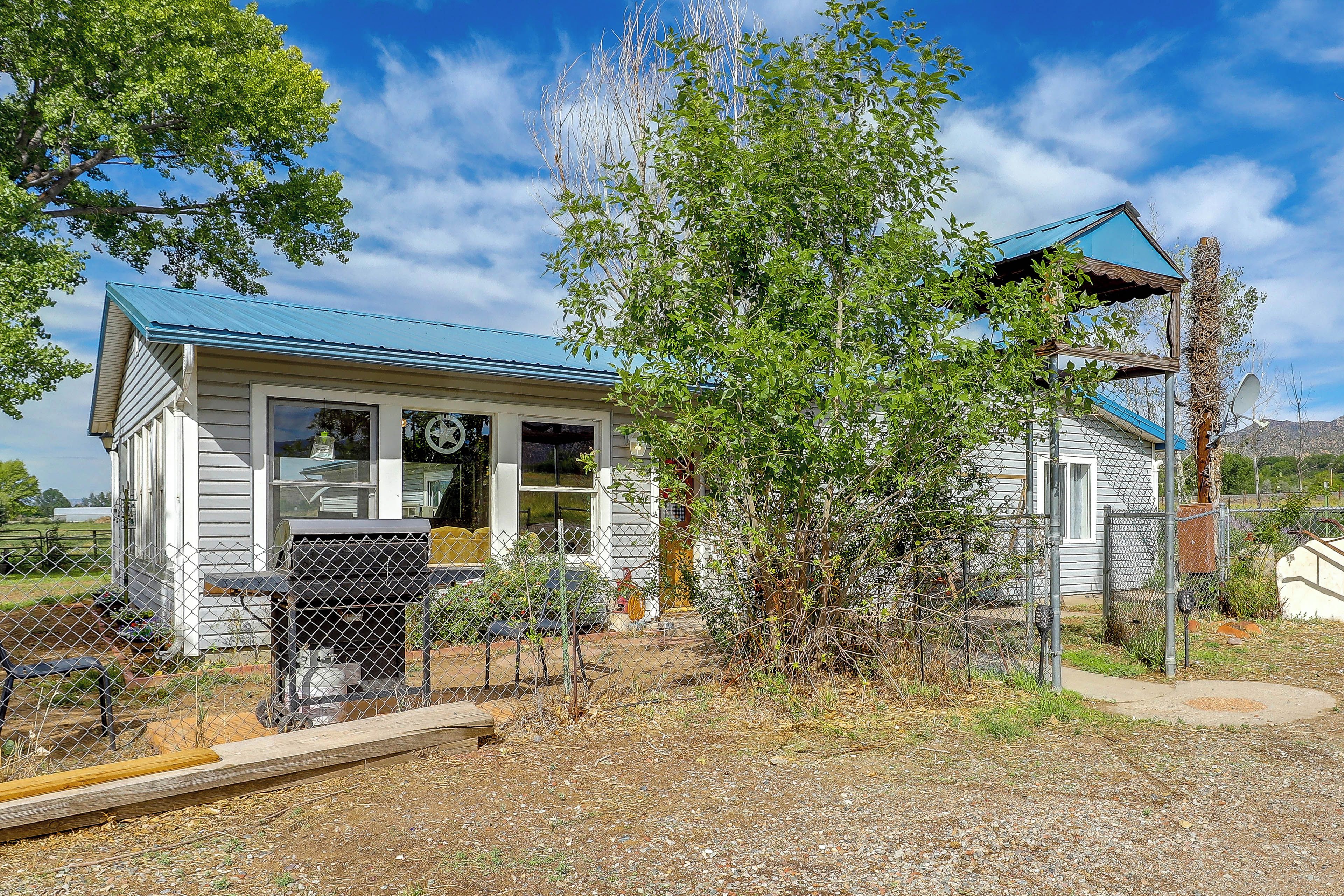 A small, single-story house with a blue roof, surrounded by a chain-link fence and various plants.
