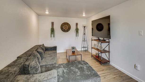 A living room with a sectional sofa, a flat-screen TV on a metal stand, a small table with a plant, and decorative wall art. - 1 Mi to Dtwn! Lubbock Home w/ Private Patio & Yard (Lubbock)