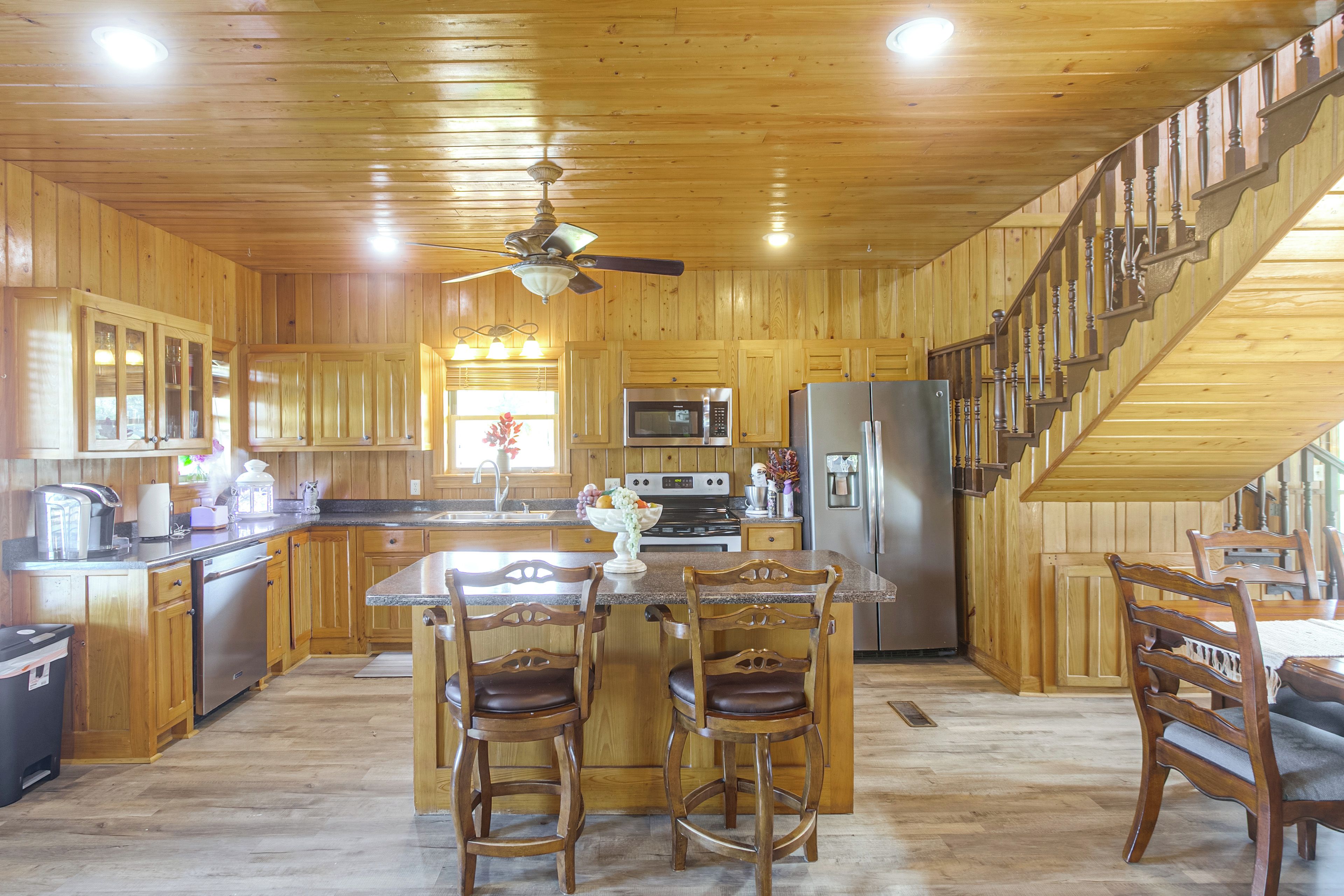 A spacious kitchen with wooden cabinets, a central island with chairs, a stainless steel refrigerator, and a staircase leading to an upper level.