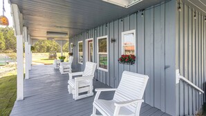 A covered porch with white chairs and a table, a vehicle in the background, and a building with vertical siding. - Georgia Cabin Escape on 500 Acres (Nashville)