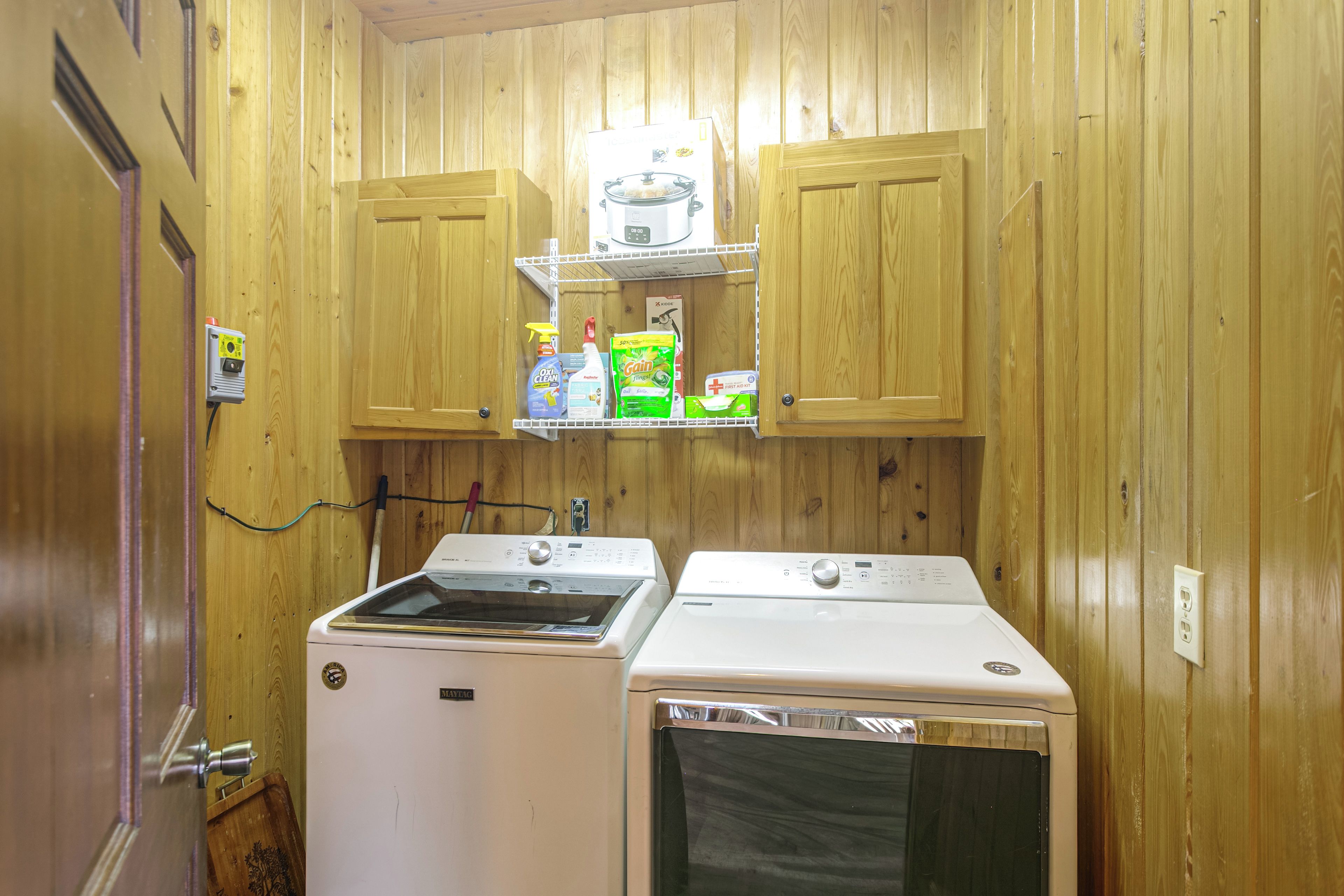 A compact laundry room with wooden walls, a washing machine, a dryer, and a shelf with cleaning supplies.