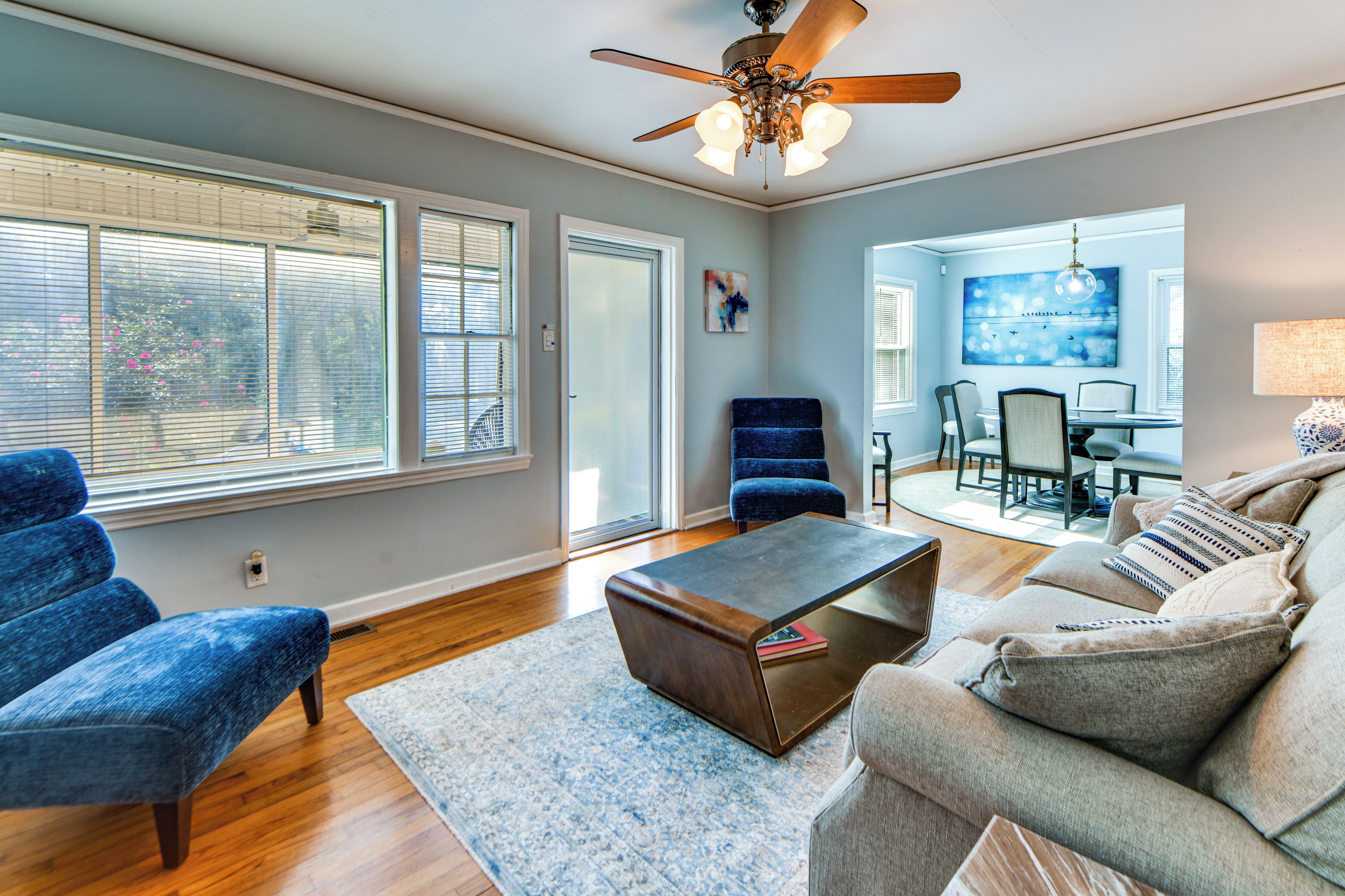 A spacious living room with a ceiling fan, a dining area with a table and chairs, and a large window with blinds.