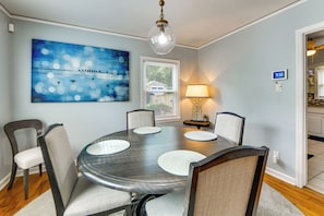 A dining room with a round table, six chairs, a painting, a window with blinds, a lamp, and a thermostat.