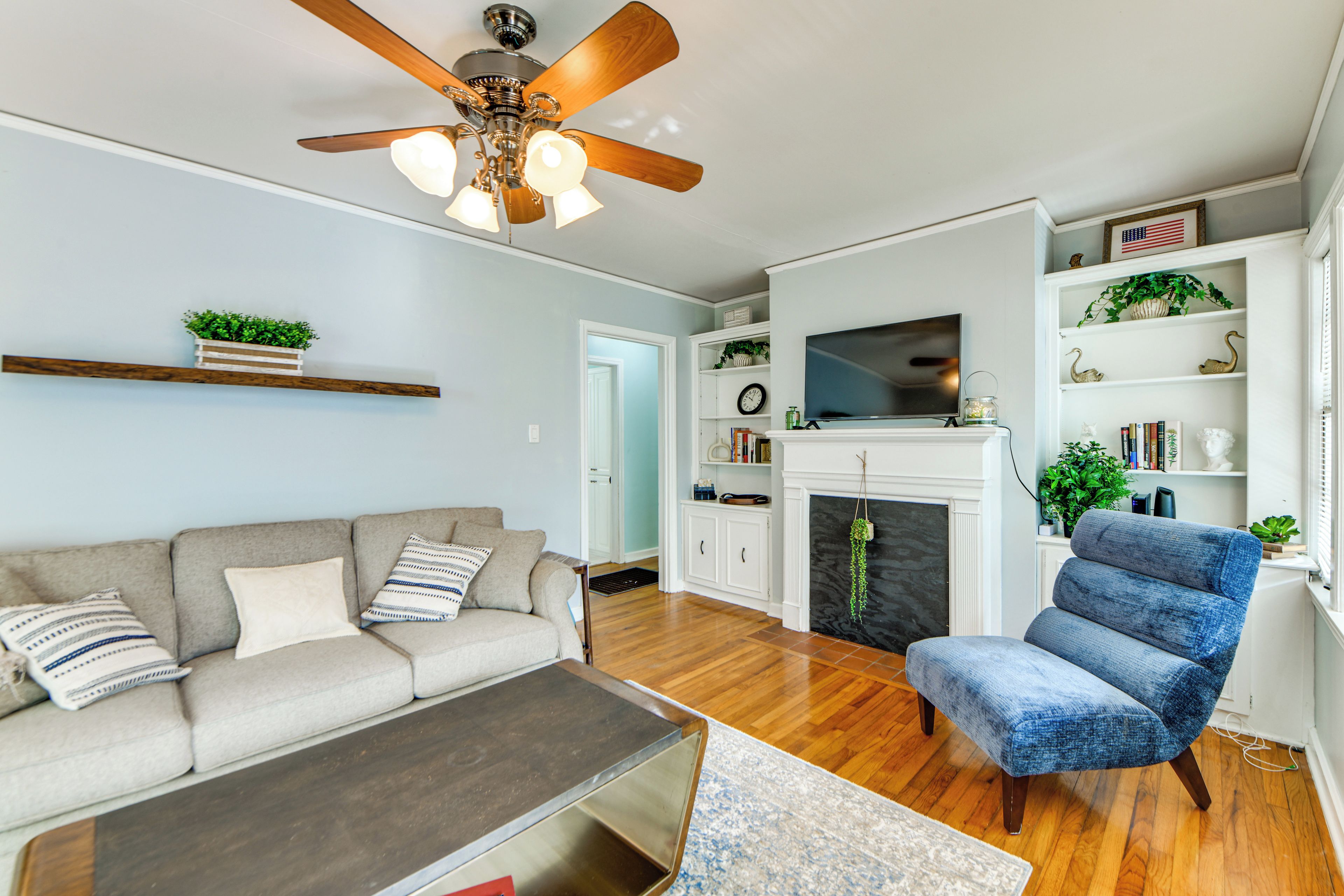 A living room with a sofa, a blue armchair, a coffee table, a fireplace, a television, and a ceiling fan.