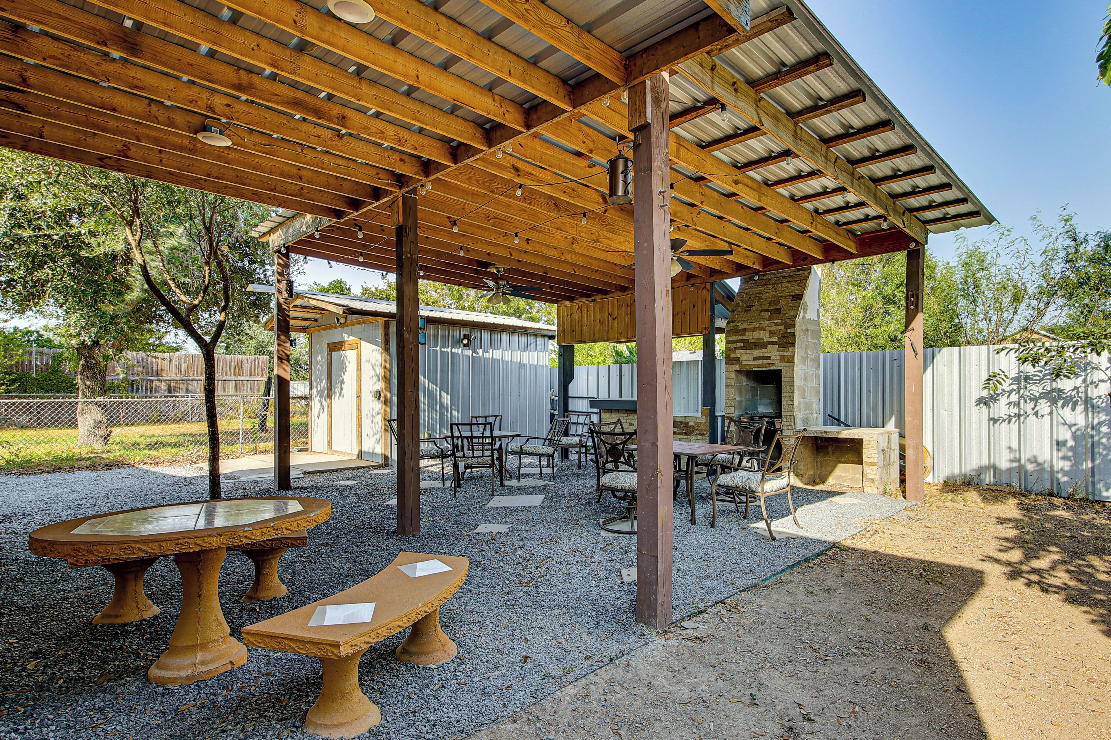 Un patio al aire libre con vigas de madera, mesas y sillas, rodeado de grava y árboles.