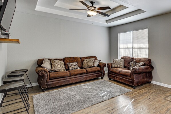 A living room with a brown leather sofa set, a ceiling fan, a window with blinds, and a wooden floor.