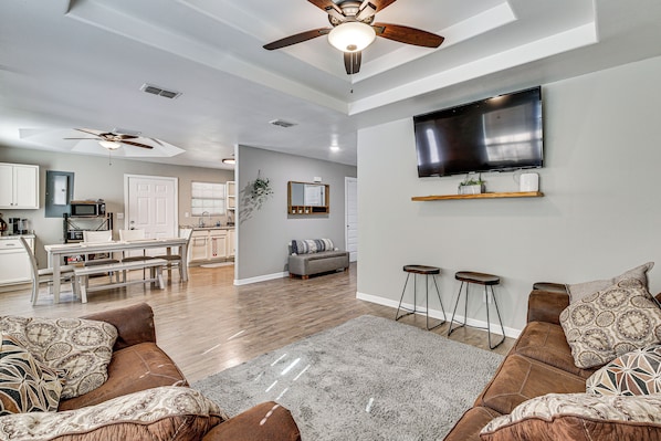 A spacious living room with a ceiling fan, a flat-screen TV mounted on the wall, a grey rug, and a seating area with patterned cushions.