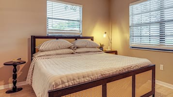 A bedroom with a bed, two windows with blinds, a bedside table with a lamp, and a small plant.