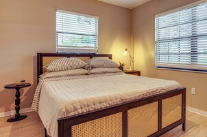 Un dormitorio con cama, dos ventanas con persianas, una mesita de noche con lámpara y una planta pequeña.
