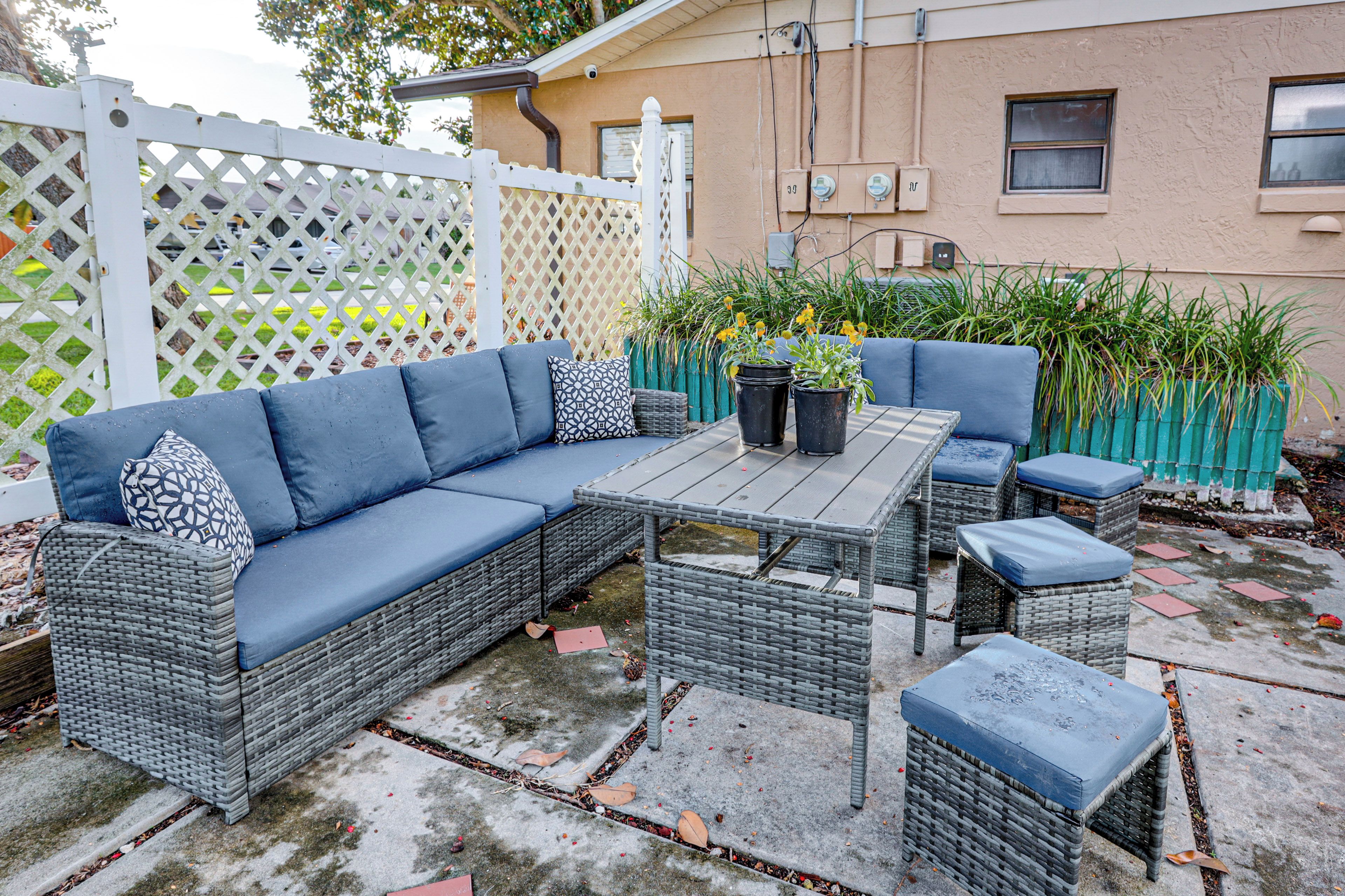 A patio area with a sectional sofa, a small table, and two chairs. There are potted plants and a white fence in the background.