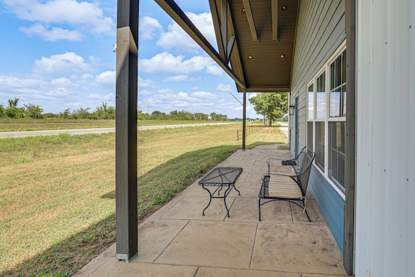 A covered patio with a concrete floor, two metal chairs, and a view of a grassy field and road.