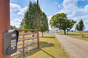 A metal gate with a keypad, a gravel path, and a fenced area with trees and a building in the background.