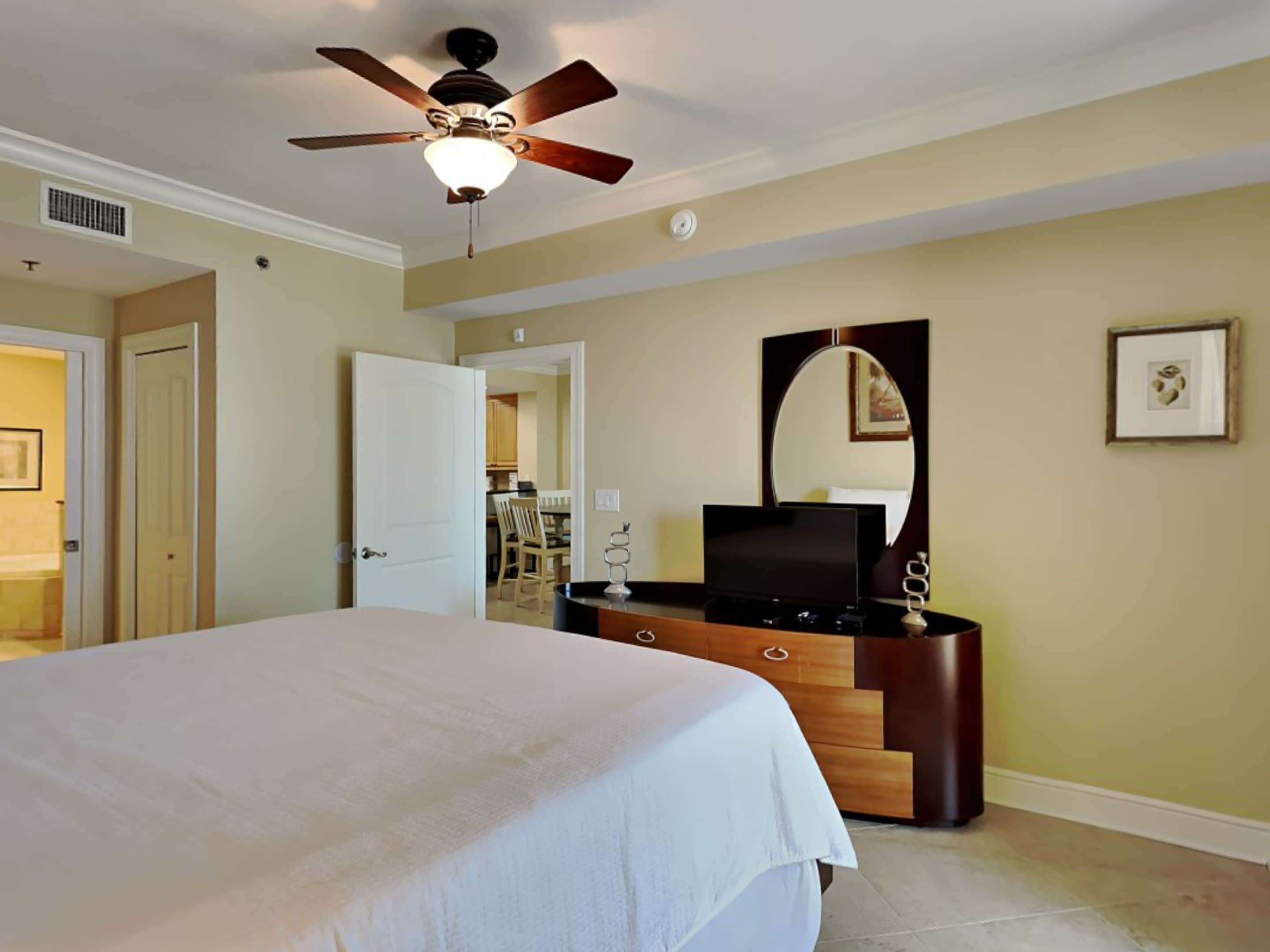 Une chambre d’hôtel comprenant un lit, une commode, un téléviseur, un ventilateur de plafond, un miroir et une porte donnant accès à une salle de bain.