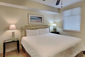 Une chambre d’hôtel avec un lit, deux tables de chevet, un ventilateur de plafond, une fenêtre avec des stores et un tableau au mur.