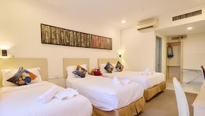 Une chambre d’hôtel avec trois lits, chacun agrémenté de draps blancs et de coussins décoratifs. Un grand tableau abstrait orne le mur au-dessus des lits.