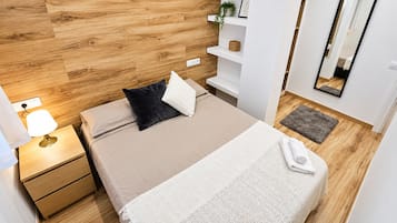 Une chambre moderne avec une tête de lit en bois, une table de chevet avec une lampe, un lit recouvert d’une couverture blanche et un petit tapis sur le sol.