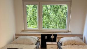 Une chambre avec deux lits, une table de chevet, une lampe et une fenêtre donnant sur des arbres.