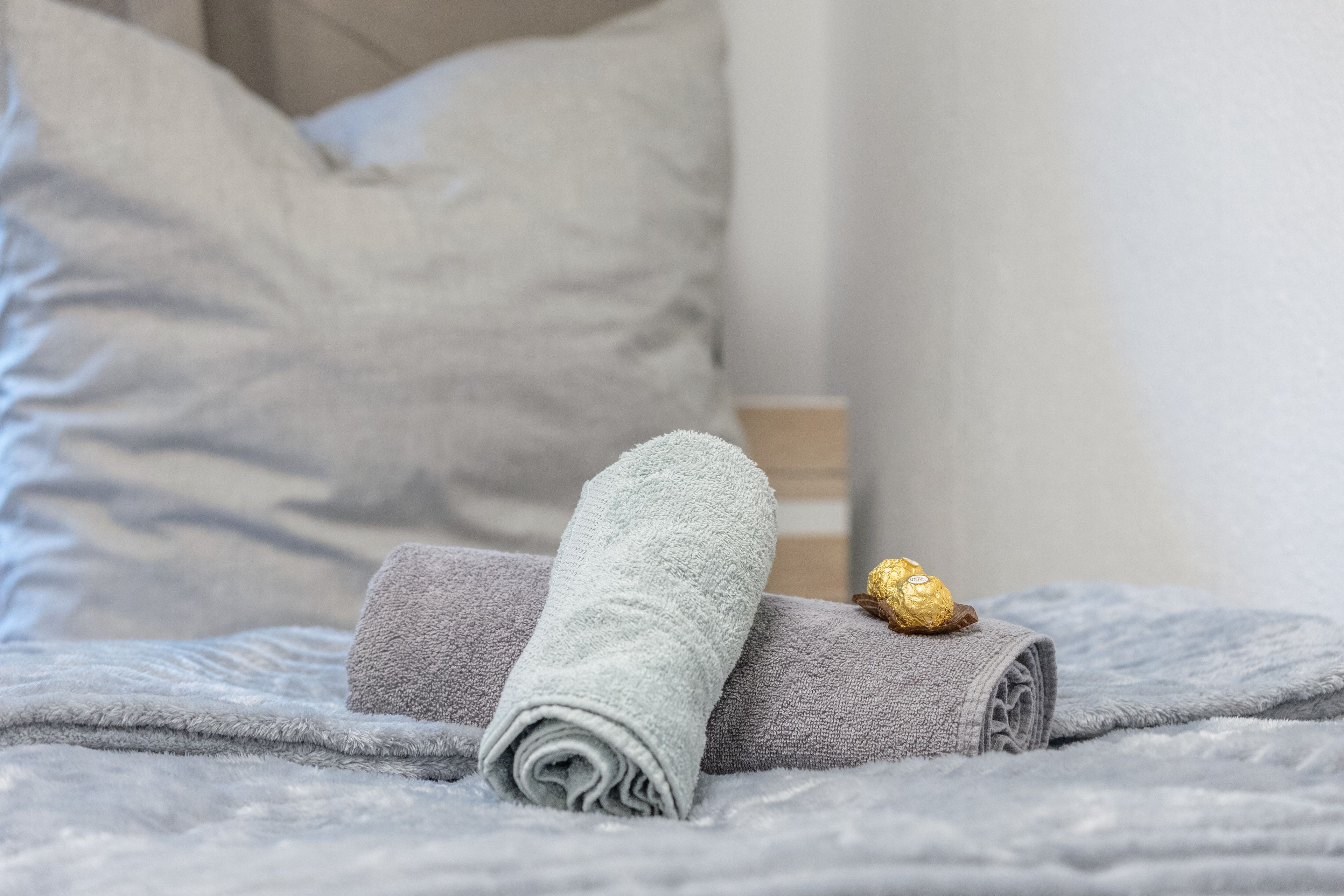 Una cama bien hecha con una toalla gris enrollada y un objeto envuelto, posiblemente una bata, sobre una manta gris con textura.