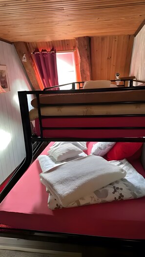 Uma cama beliche com colchão vermelho e almofadas brancas, paredes de madeira e uma janela com cortinas.