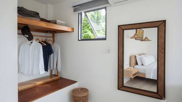 A compact hotel room with a wooden bench, a mirror, a small window, and a wall-mounted air conditioner.