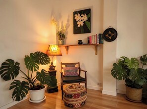 A cozy room with a wooden floor, a chair with a patterned cushion, a table lamp, and large potted plants.