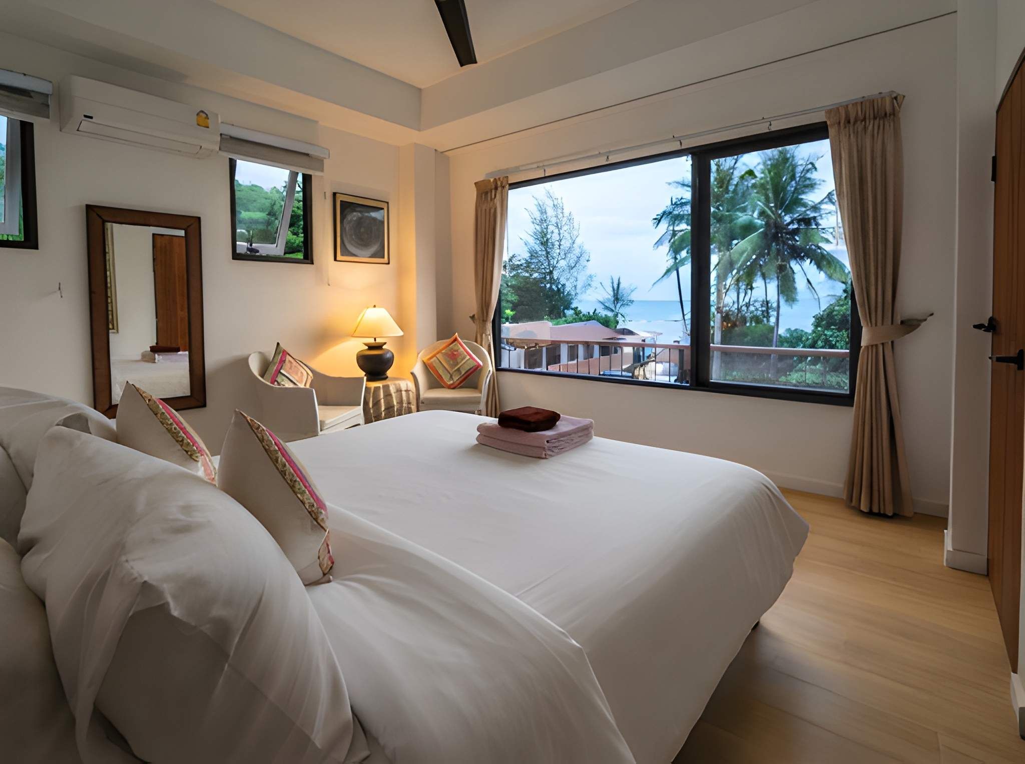 A hotel room with a large bed, a desk with a lamp, a window with a view of palm trees, and a door.