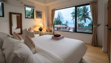 A hotel room with a large bed, a desk with a lamp, a window with a view of palm trees, and a door.