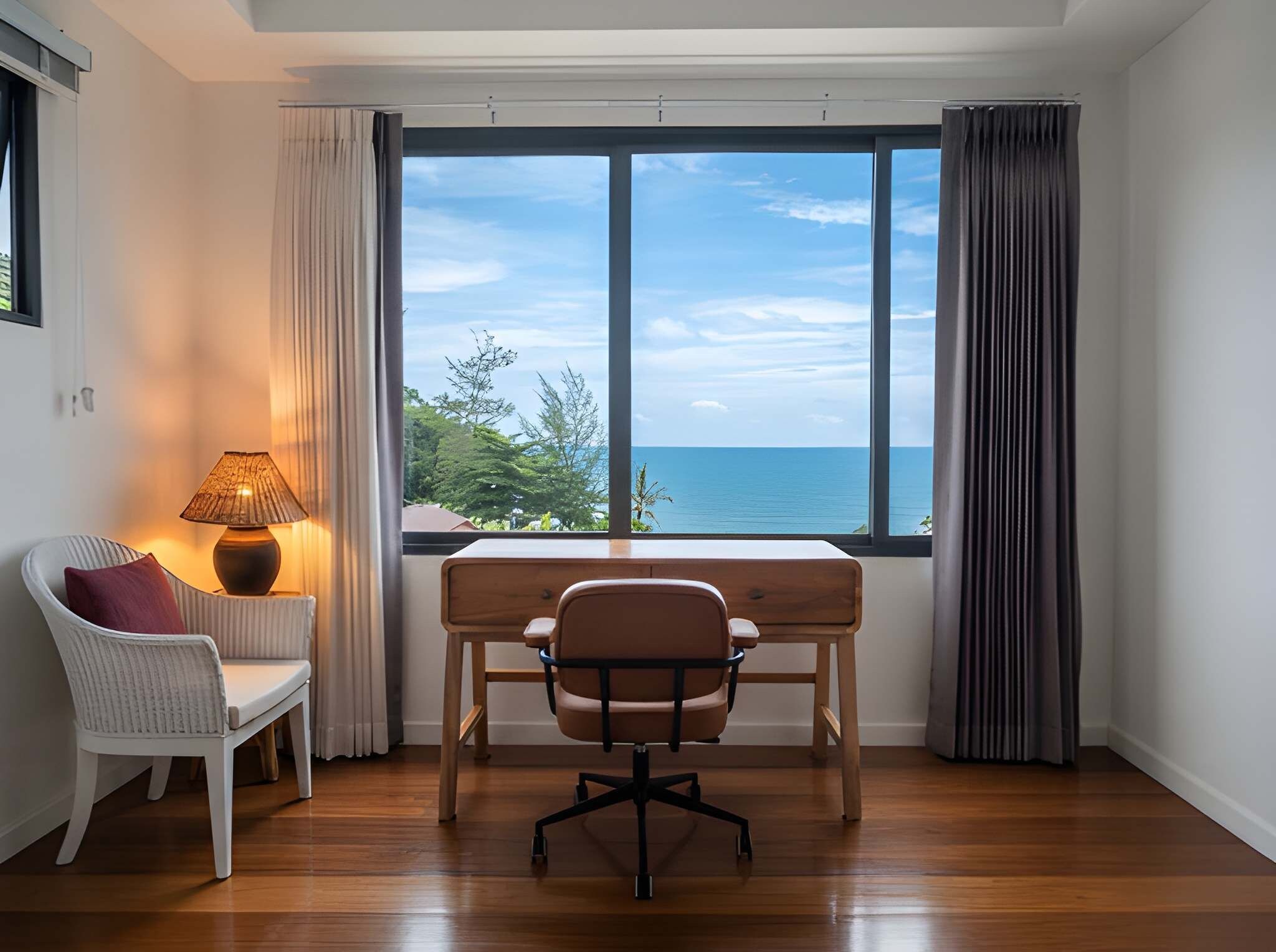 Ein Zimmer mit Schreibtisch aus Holz, einem Stuhl, einer Lampe und Meerblick durch große Fenster.