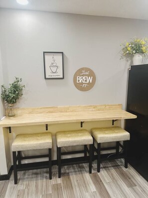 A kitchen area with a wooden countertop, two stools, a framed picture, a round sign, and a potted plant.