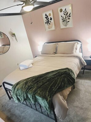Ein ordentlich bezogenes Bett mit grüner Decke, ein Nachttisch mit Lampe und Wandbild über dem Bett.