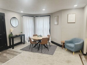 Ein Zimmer mit einem runden Tisch, Stühlen, einem blauen Sessel, einem kleinen Beistelltisch und einem Spiegel an der Wand.