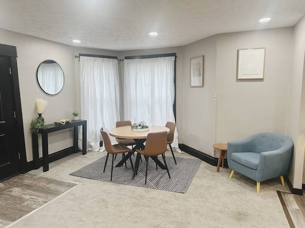 A room with a round table, chairs, a blue armchair, a small side table, and a mirror on the wall.