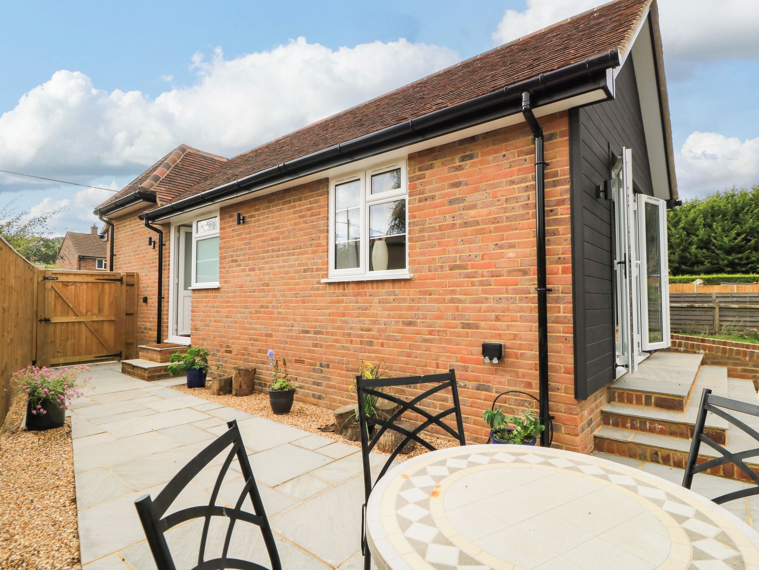 A brick house with a tiled patio, a wooden gate, and a small outdoor seating area with chairs.