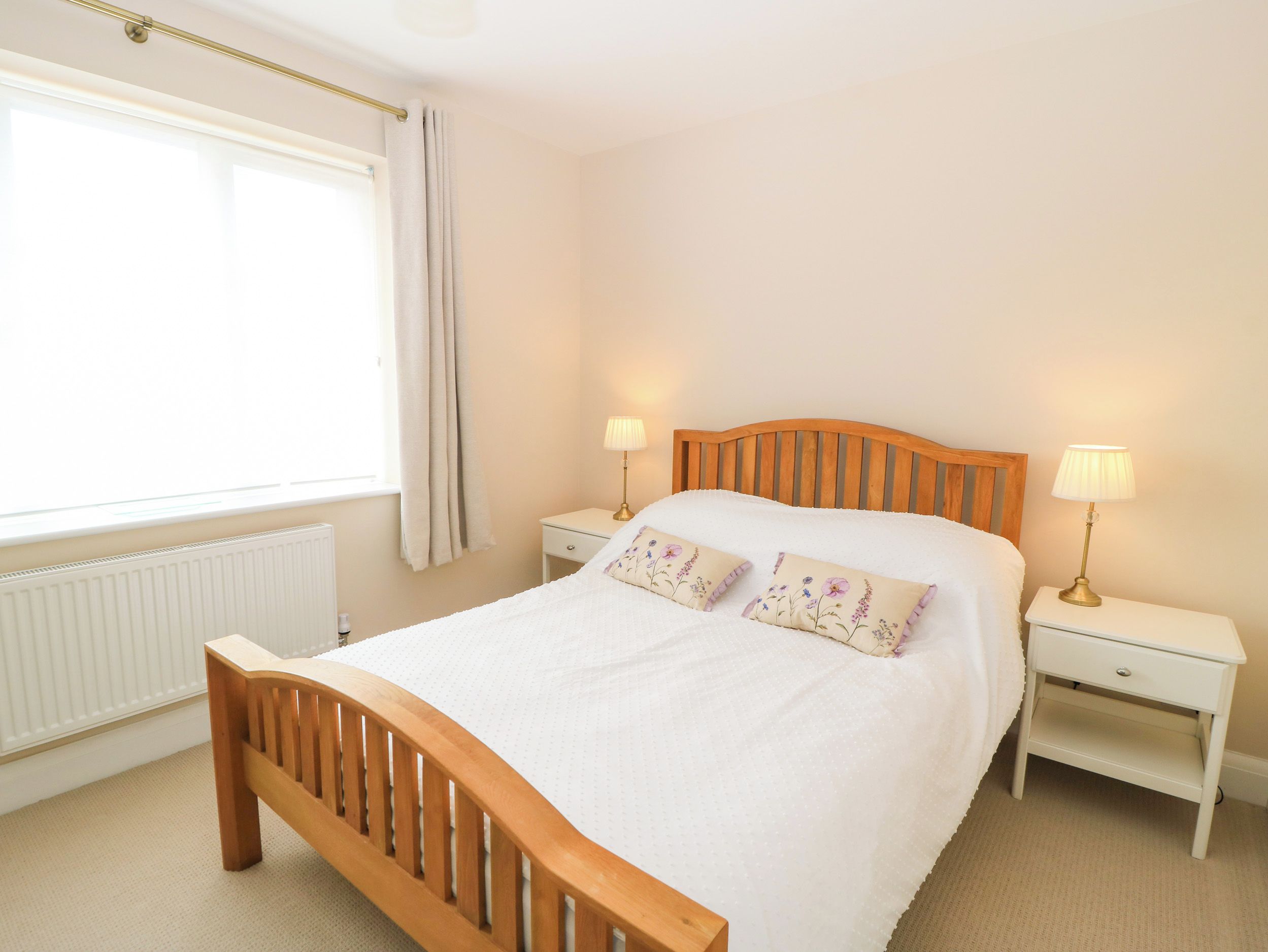 A bedroom with a wooden bed, two bedside tables with lamps, a window with curtains, and a radiator.