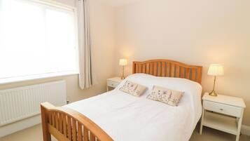 A bedroom with a wooden bed, two bedside tables with lamps, a window with curtains, and a radiator.