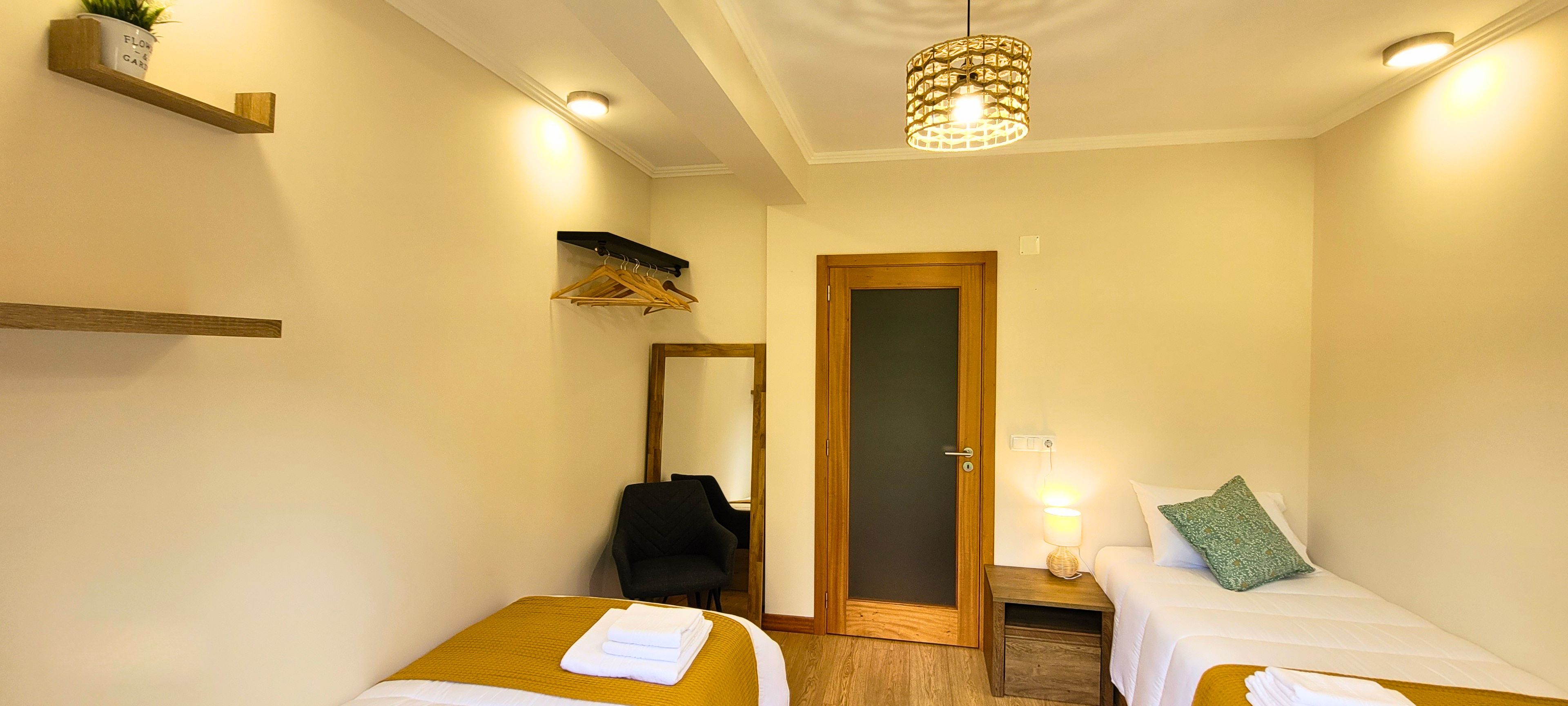A hotel room with two beds, a wooden door, a mirror, a hanging light fixture, and a small bedside table with a lamp.
