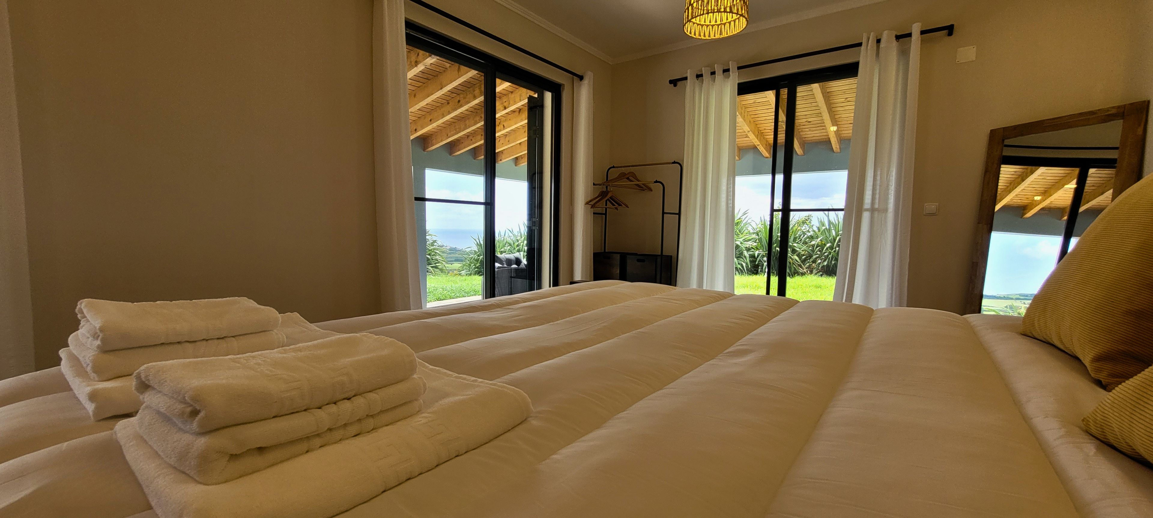 A hotel room with a large bed, two windows, a view of greenery, and a hanging light fixture.