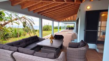 Een overdekt terras met een bank, stoelen en een tafel, met uitzicht op een weelderige tuin en de zee.