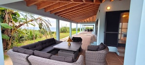 A covered outdoor seating area with a sofa, chairs, and a table, overlooking a lush garden and the sea.