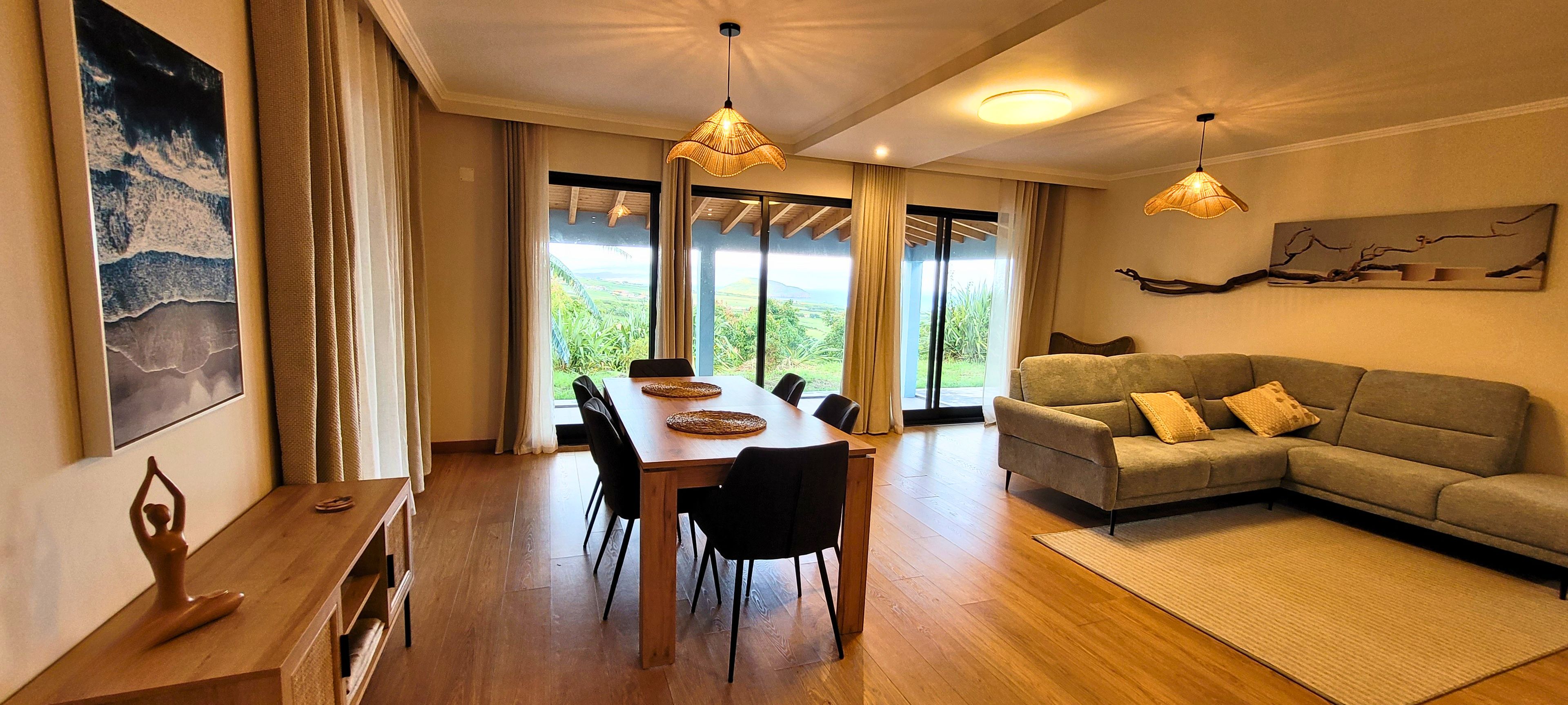 A spacious room with a wooden dining table, black chairs, a grey sofa, and a large window offering a view of greenery.
