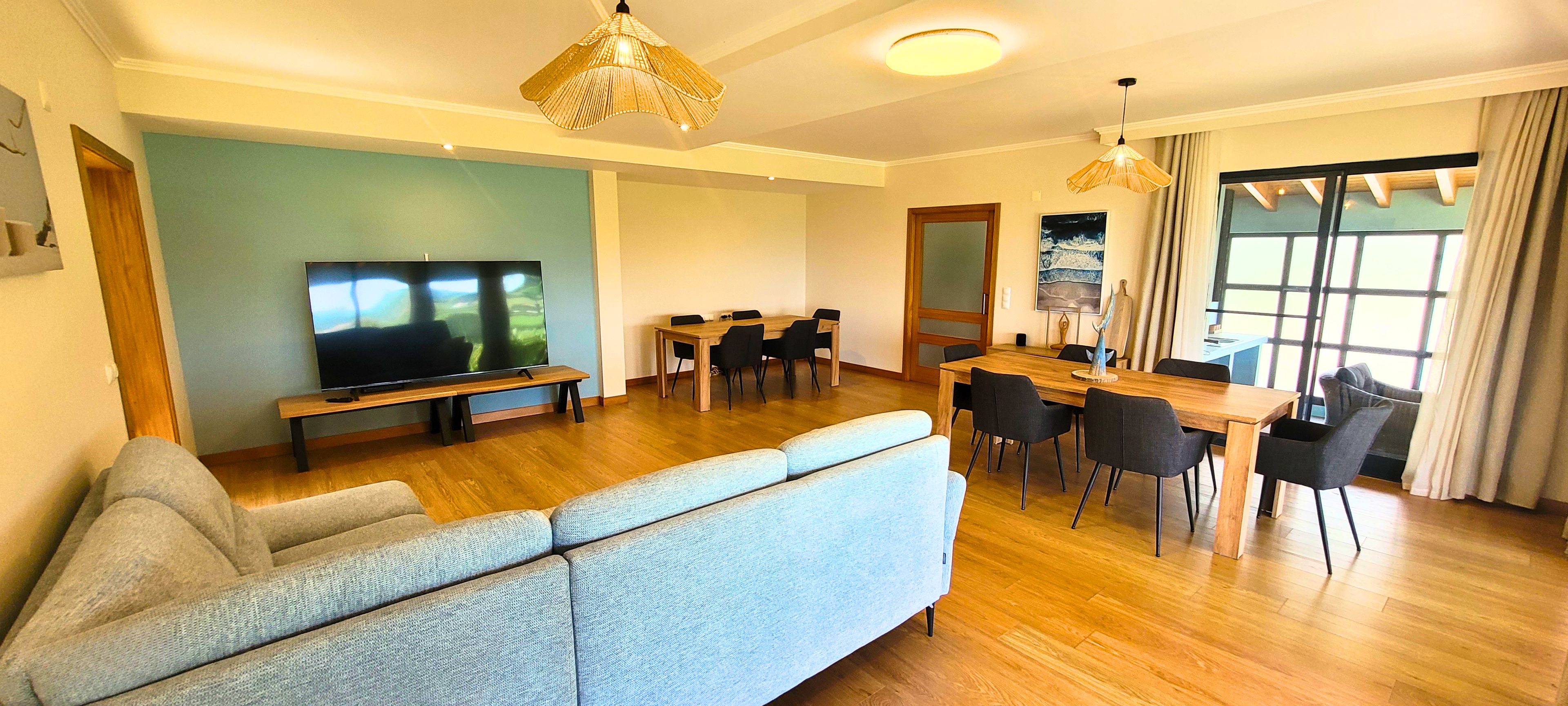 A modern living room with a large flat-screen TV, a sofa, a dining area with a wooden table and chairs, and a large window with curtains.