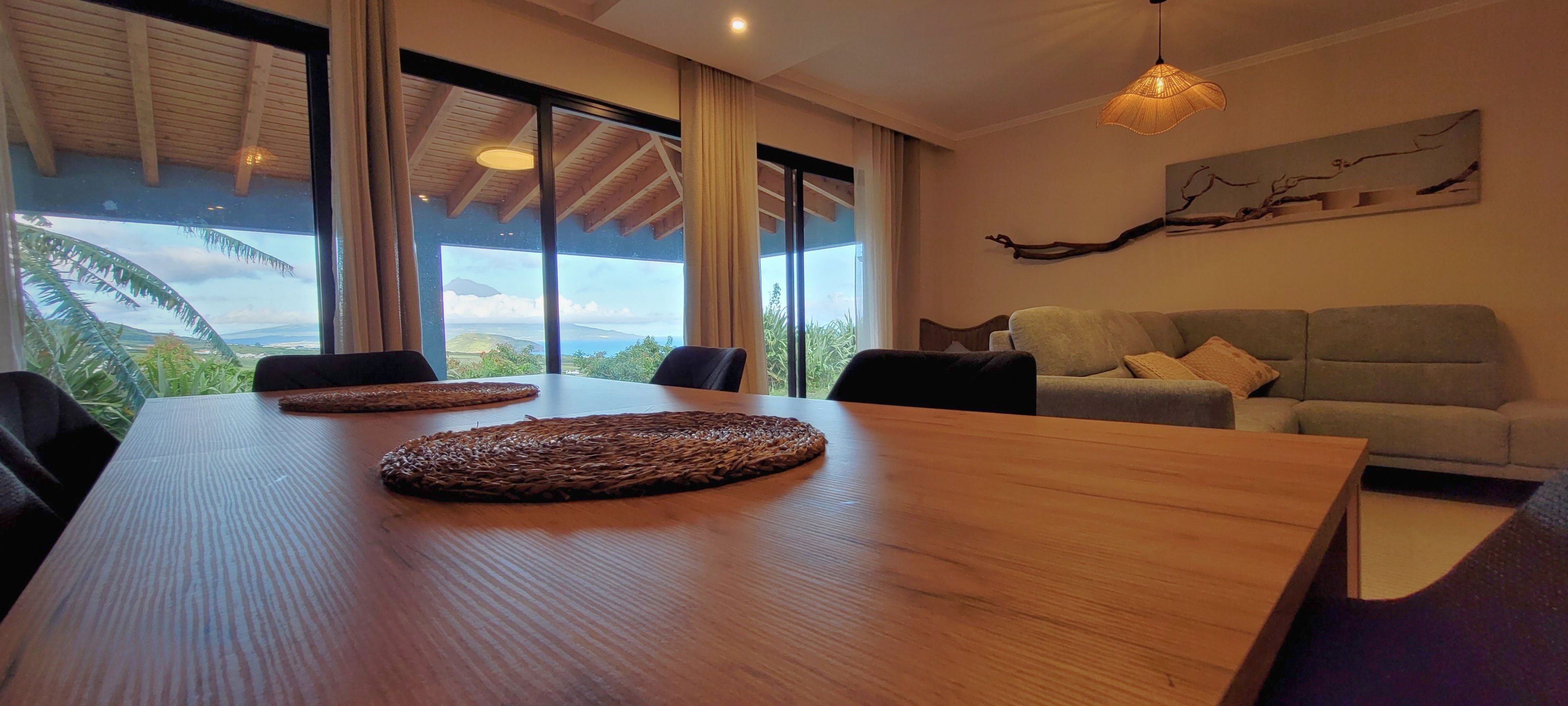 A modern living room with a wooden dining table, a grey sofa, and a large window offering a view of the outdoors.