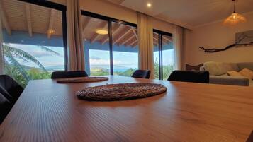 A modern living room with a wooden dining table, a grey sofa, and a large window offering a view of the outdoors.