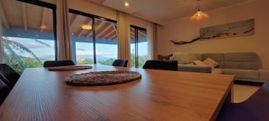 A modern living room with a wooden dining table, a grey sofa, and a large window offering a view of the outdoors.