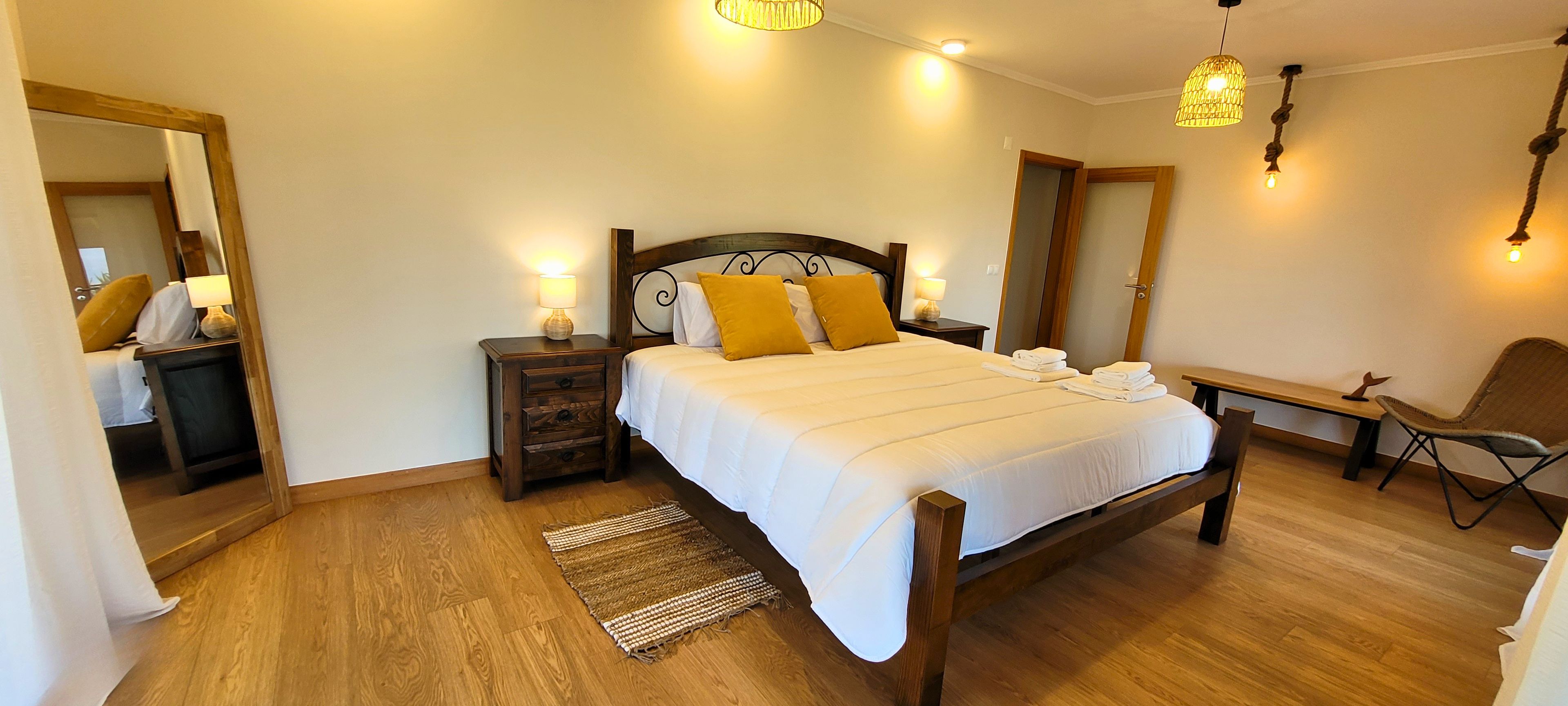 Een goed verlichte slaapkamer met een houten bed, nachtkastjes met lampen, een bankje en een stoel.