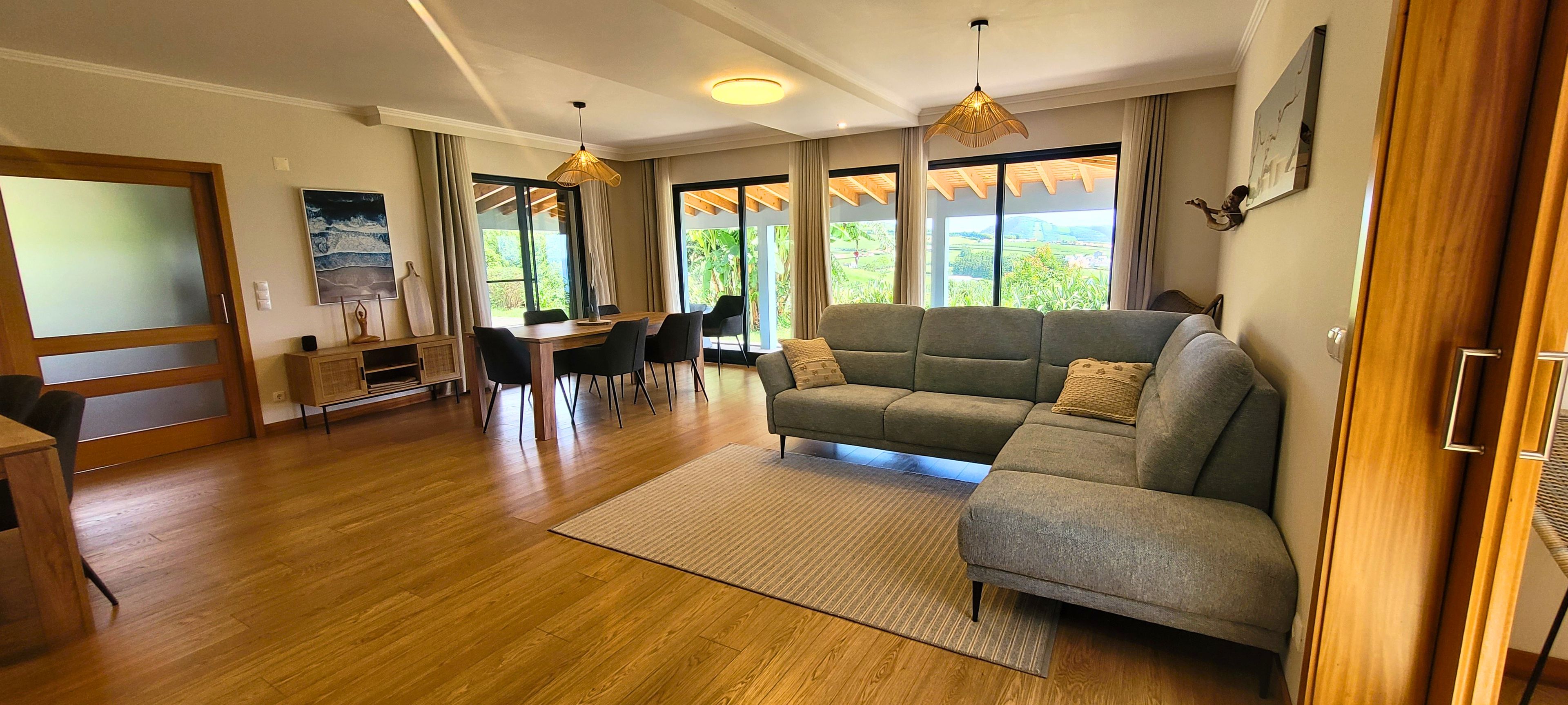 Ein Wohnzimmer mit einer Couch, einem Esstisch mit Stühlen, einem Fernseher und großen Fenstern mit Vorhängen.