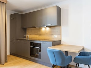 A modern kitchen with grey cabinets, a wooden backsplash, and a built-in oven. There are two blue chairs at a wooden table. - Studio for 2 People (Mittersill)