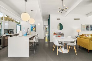 A modern kitchen with a central island, white dining chairs, a yellow sofa, and a dining table. - Janet’s Hideaway — Classic 1961 Alexander with Private Pool & Spa (Palm Springs)