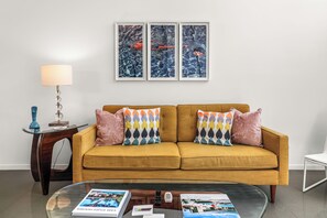 A living room with a mustard-colored sofa, two patterned throw pillows, a glass coffee table with magazines, a lamp, and three framed aquatic paintings on the wall. - Janet’s Hideaway — Classic 1961 Alexander with Private Pool & Spa (Palm Springs)