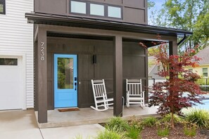 Ein modernes, zweistöckiges Haus mit einer blauen Tür, zwei weißen Schaukelstühlen und einem kleinen Baum mit roten Blättern.